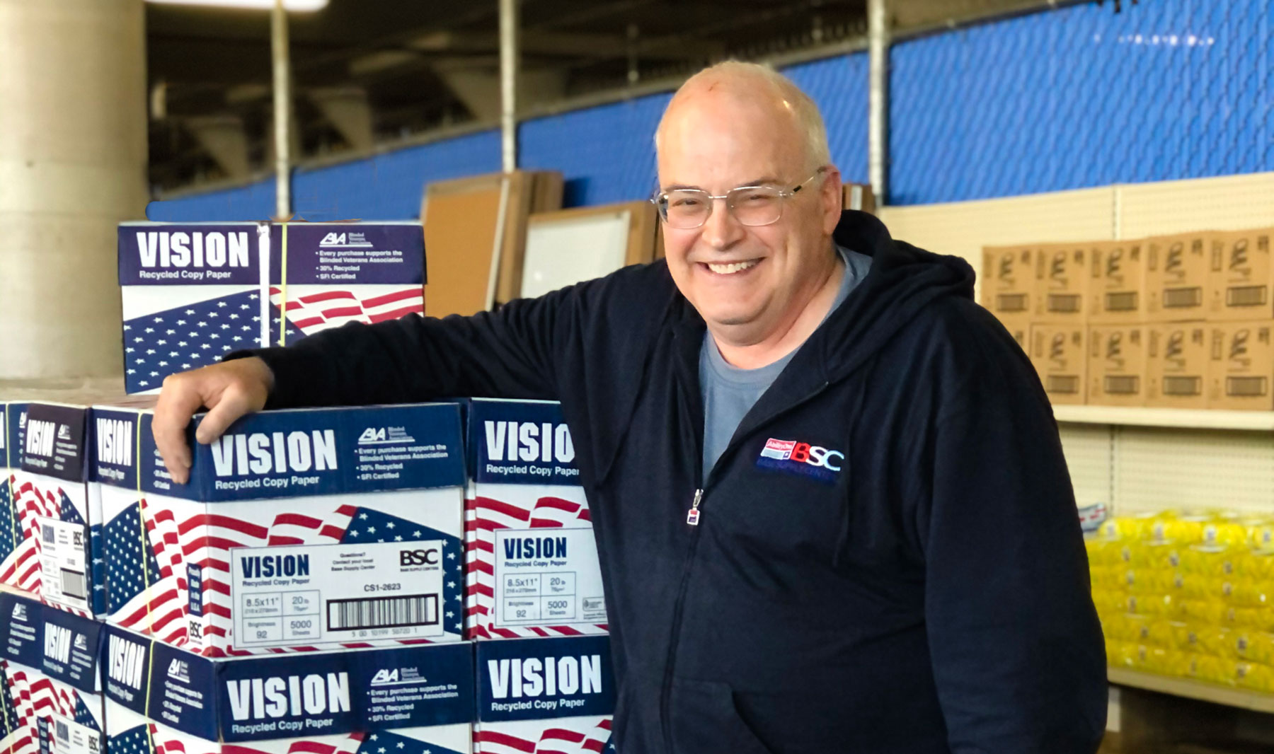 Image Description: A middle aged white man leans on a tower of stacked boxes, smiling. He is wearing glasses and a dark blue sweatshirt with an embroidered BSC logo.